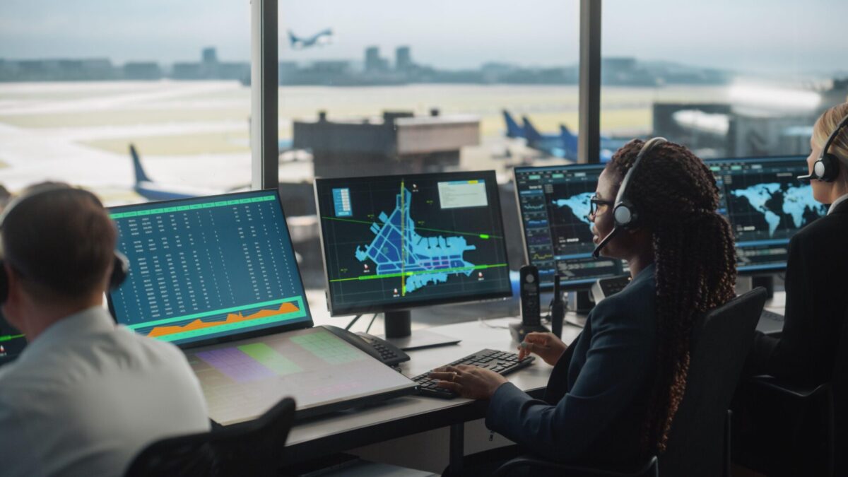May 2nd Edition Article 4 Team of airport operations staff wearing headsets monitor multiple screens showing flight data and a control room map with the runway visible outside the window