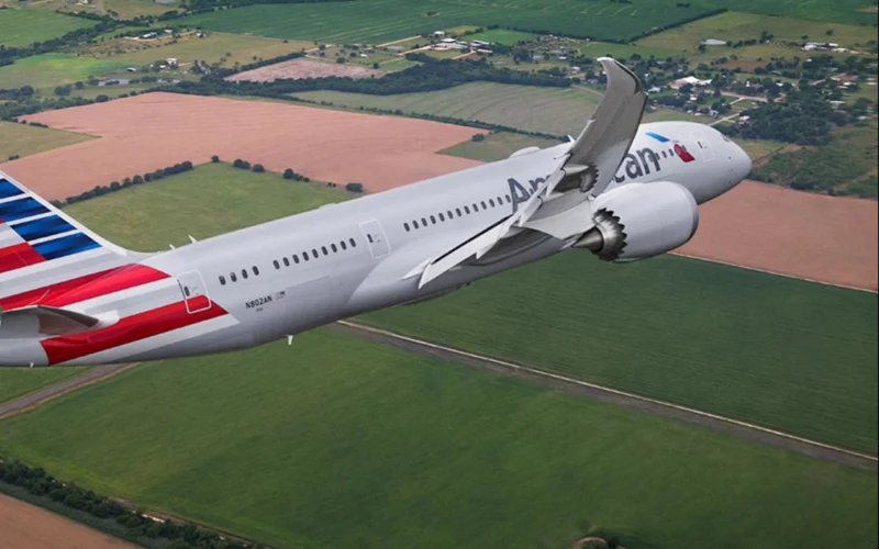 Commercial airliner in flight over green and brown fields with American Airlines livery visible on the fuselage and tailfin
