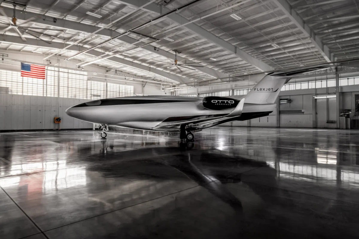 Inside a spacious hangar a sleek white and black Otto Aerospace aircraft with Flexjet branding sits on the polished floor with an American flag visible on the wall overhead