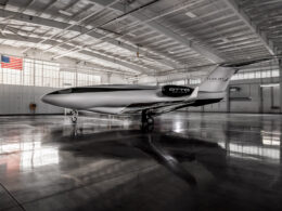 Inside a spacious hangar a sleek white and black Otto Aerospace aircraft with Flexjet branding sits on the polished floor with an American flag visible on the wall overhead