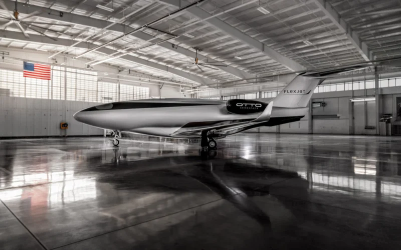 Inside a spacious hangar a sleek white and black Otto Aerospace aircraft with Flexjet branding sits on the polished floor with an American flag visible on the wall overhead