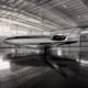 Inside a spacious hangar a sleek white and black Otto Aerospace aircraft with Flexjet branding sits on the polished floor with an American flag visible on the wall overhead