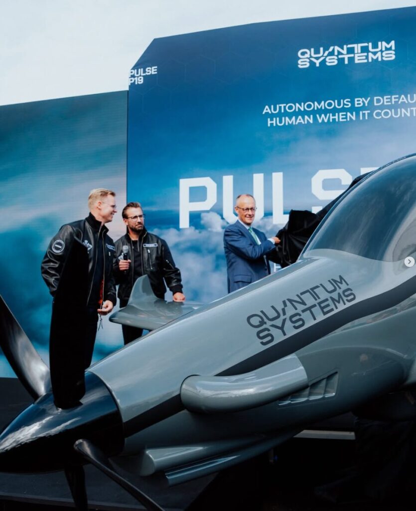 Screenshot 2026 06 10 132533 Three men stand beside a gray prototype aircraft labeled QUANTUM SYSTEMS at a blue event backdrop reading PULSE 2019