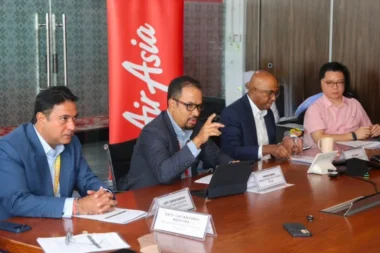 Four professionals in a boardroom at a conference table one man in a suit is speaking while others listen nameplates in front of them