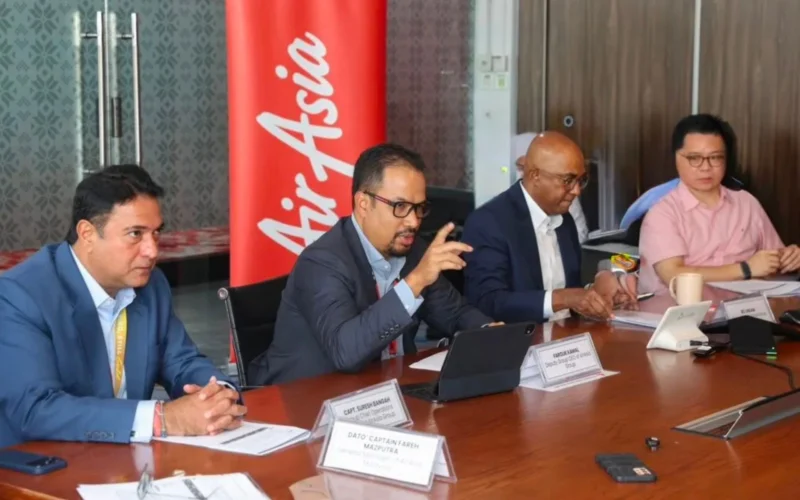 Four professionals in a boardroom at a conference table one man in a suit is speaking while others listen nameplates in front of them