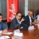 Four professionals in a boardroom at a conference table one man in a suit is speaking while others listen nameplates in front of them
