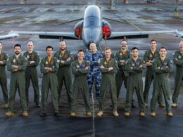 Group of pilots in green flight suits posing with arms crossed in front of a jet on a taxiway runway