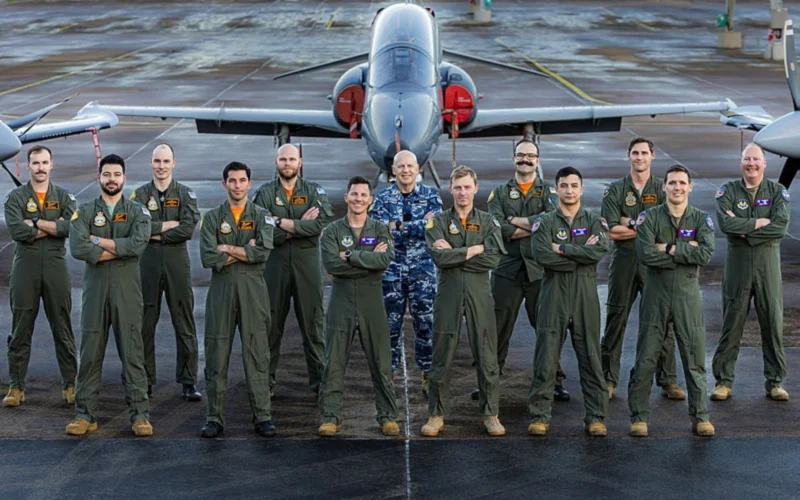 Group of pilots in green flight suits posing with arms crossed in front of a jet on a taxiway runway