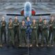 Group of pilots in green flight suits posing with arms crossed in front of a jet on a taxiway runway