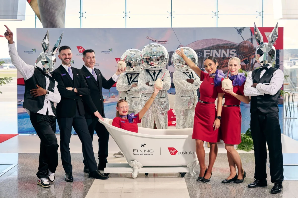 Virgin Australia flies first international Canberra flight Group of staff and performers pose around a white bathtub with cocktail drinks at a promo event some wearing silver mirrored bunny heads and others in red dresses and black suits backdrop shows sponsors