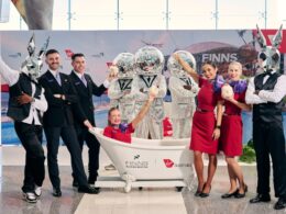 Group of staff and performers pose around a white bathtub with cocktail drinks at a promo event some wearing silver mirrored bunny heads and others in red dresses and black suits backdrop shows sponsors