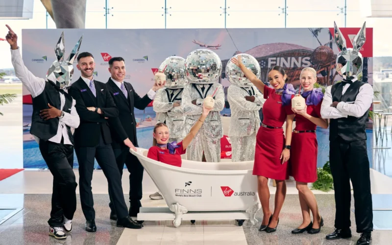 Group of staff and performers pose around a white bathtub with cocktail drinks at a promo event some wearing silver mirrored bunny heads and others in red dresses and black suits backdrop shows sponsors