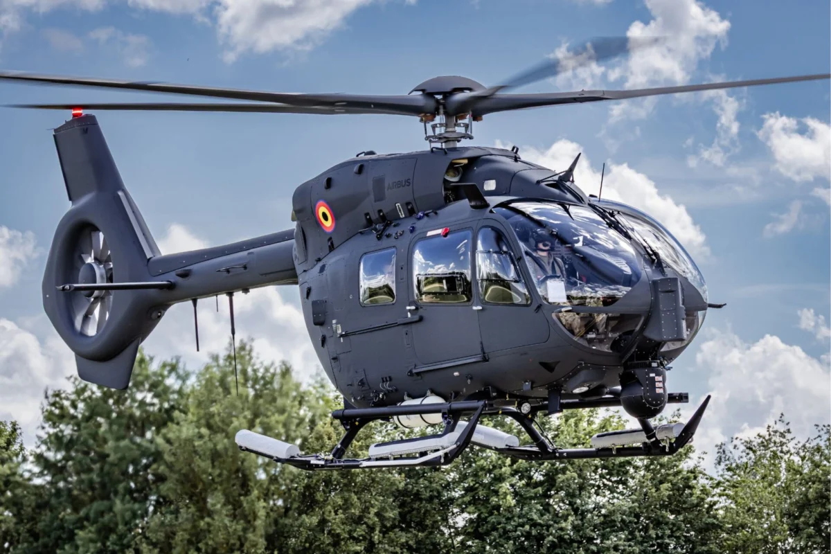 Gray Airbus helicopter hovering in the air with spinning rotor blades cockpit windows visible against a cloudy blue sky and trees below