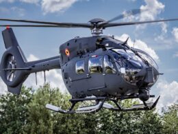 Gray Airbus helicopter hovering in the air with spinning rotor blades cockpit windows visible against a cloudy blue sky and trees below