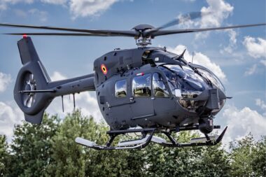 Gray Airbus helicopter hovering in the air with spinning rotor blades cockpit windows visible against a cloudy blue sky and trees below