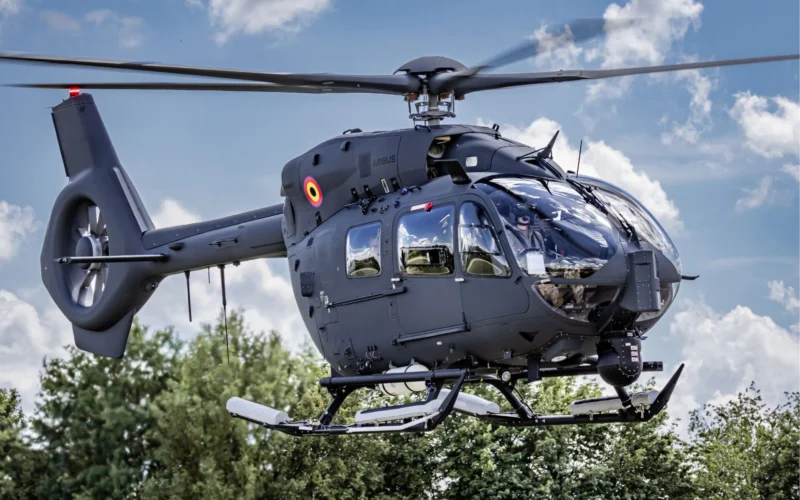 Gray Airbus helicopter hovering in the air with spinning rotor blades cockpit windows visible against a cloudy blue sky and trees below