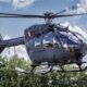 Gray Airbus helicopter hovering in the air with spinning rotor blades cockpit windows visible against a cloudy blue sky and trees below