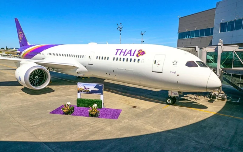 Thai Airways aircraft docked at a gate with a purple tail and logo boarding bridge on the right and a purple floral display in the foreground