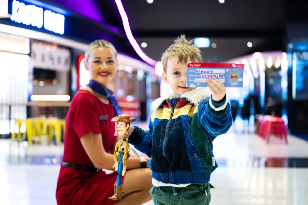 A young boy in a colorful jacket holds a Toy Story Woody figurine and an admission ticket with a smiling staff member in a red uniform nearby in a bright venue