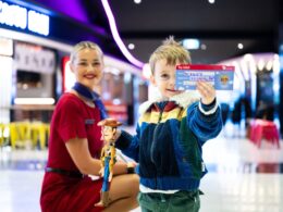 A young boy in a colorful jacket holds a Toy Story Woody figurine and an admission ticket with a smiling staff member in a red uniform nearby in a bright venue