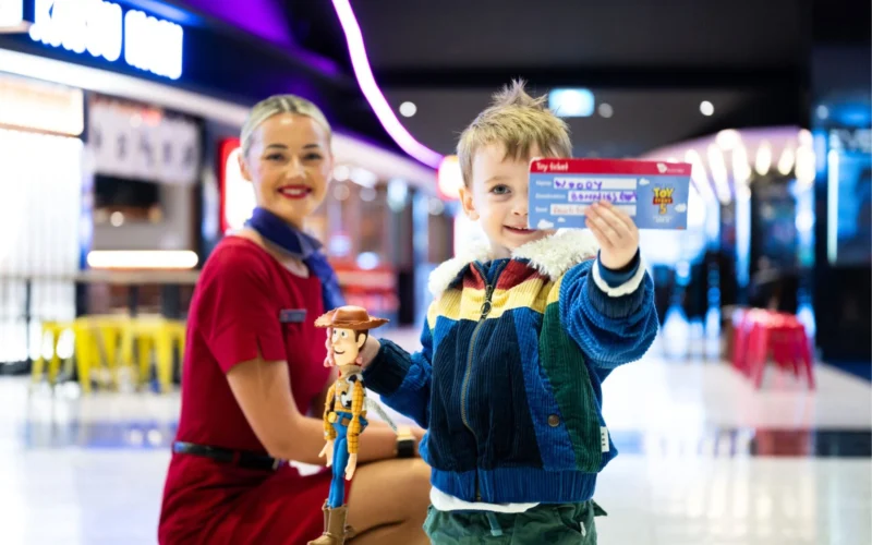A young boy in a colorful jacket holds a Toy Story Woody figurine and an admission ticket with a smiling staff member in a red uniform nearby in a bright venue