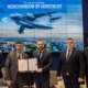 Five men in suits pose with a signed memorandum of agreement in front of a large blue backdrop displaying a cargo aircraft and a city skyline