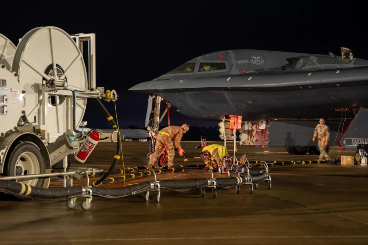 Nighttime on an airfield a military fighter jet is being serviced by ground crew with hoses and equipment nearby