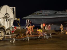 Nighttime on an airfield a military fighter jet is being serviced by ground crew with hoses and equipment nearby