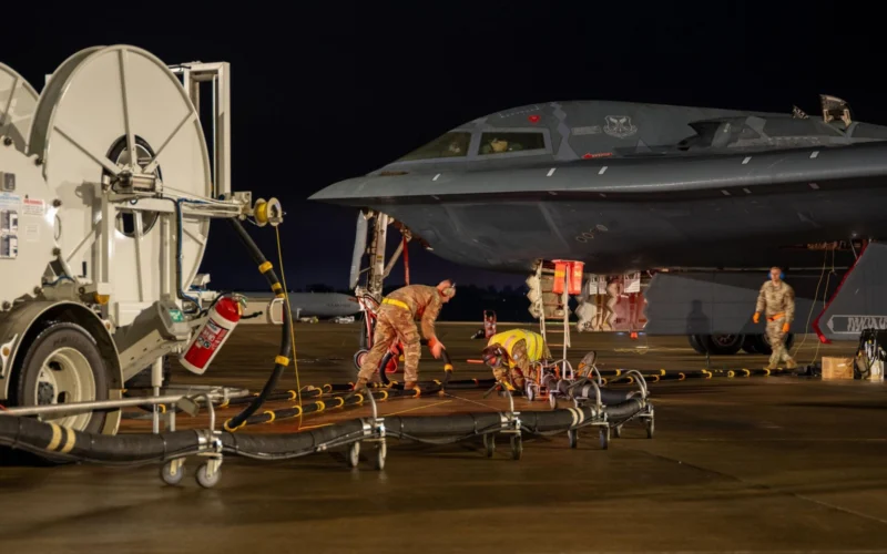 Nighttime on an airfield a military fighter jet is being serviced by ground crew with hoses and equipment nearby