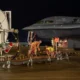 Nighttime on an airfield a military fighter jet is being serviced by ground crew with hoses and equipment nearby