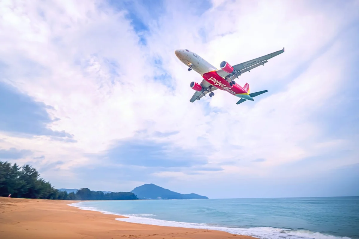 Red VietJet airliner in flight above a sandy beach and calm blue sea with distant hills on the horizon