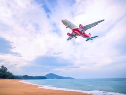 Red VietJet airliner in flight above a sandy beach and calm blue sea with distant hills on the horizon