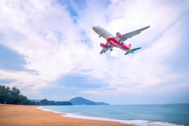 Red VietJet airliner in flight above a sandy beach and calm blue sea with distant hills on the horizon