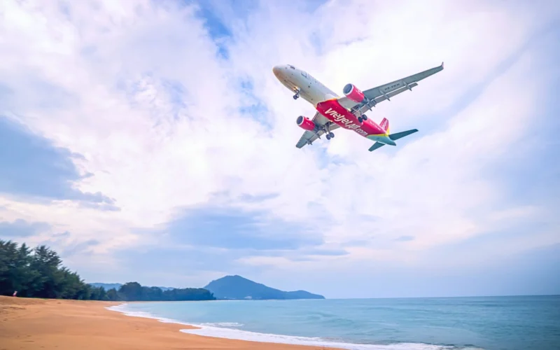 Red VietJet airliner in flight above a sandy beach and calm blue sea with distant hills on the horizon