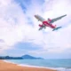 Red VietJet airliner in flight above a sandy beach and calm blue sea with distant hills on the horizon