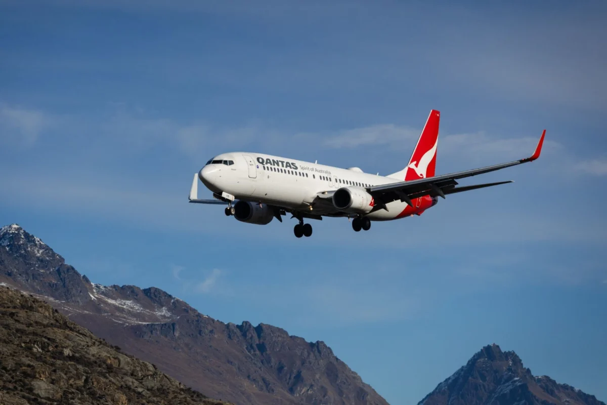 Qantas passenger jet in flight over rugged mountain landscape with a clear blue sky tail logo visible