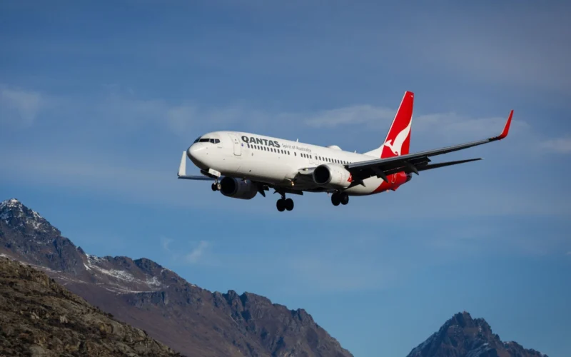 Qantas passenger jet in flight over rugged mountain landscape with a clear blue sky tail logo visible