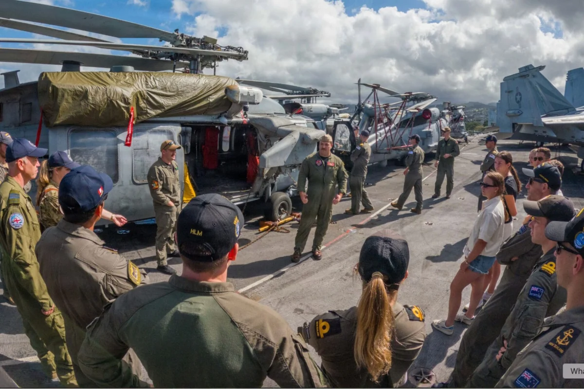 Crew members in flight suits stand on an aircraft carrier deck beside parked helicopters and aircraft structures in the background