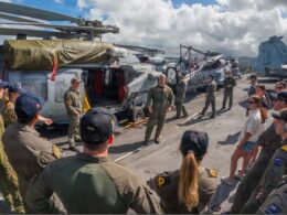 Crew members in flight suits stand on an aircraft carrier deck beside parked helicopters and aircraft structures in the background