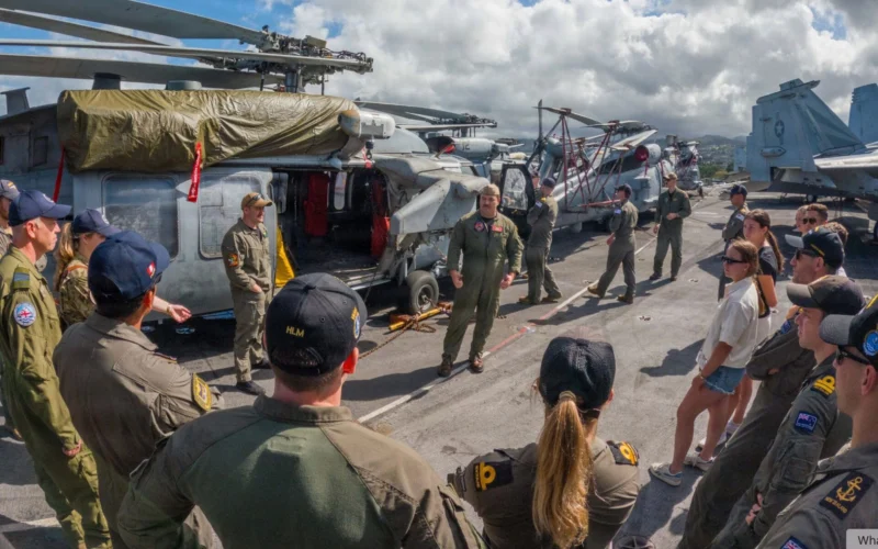 Crew members in flight suits stand on an aircraft carrier deck beside parked helicopters and aircraft structures in the background
