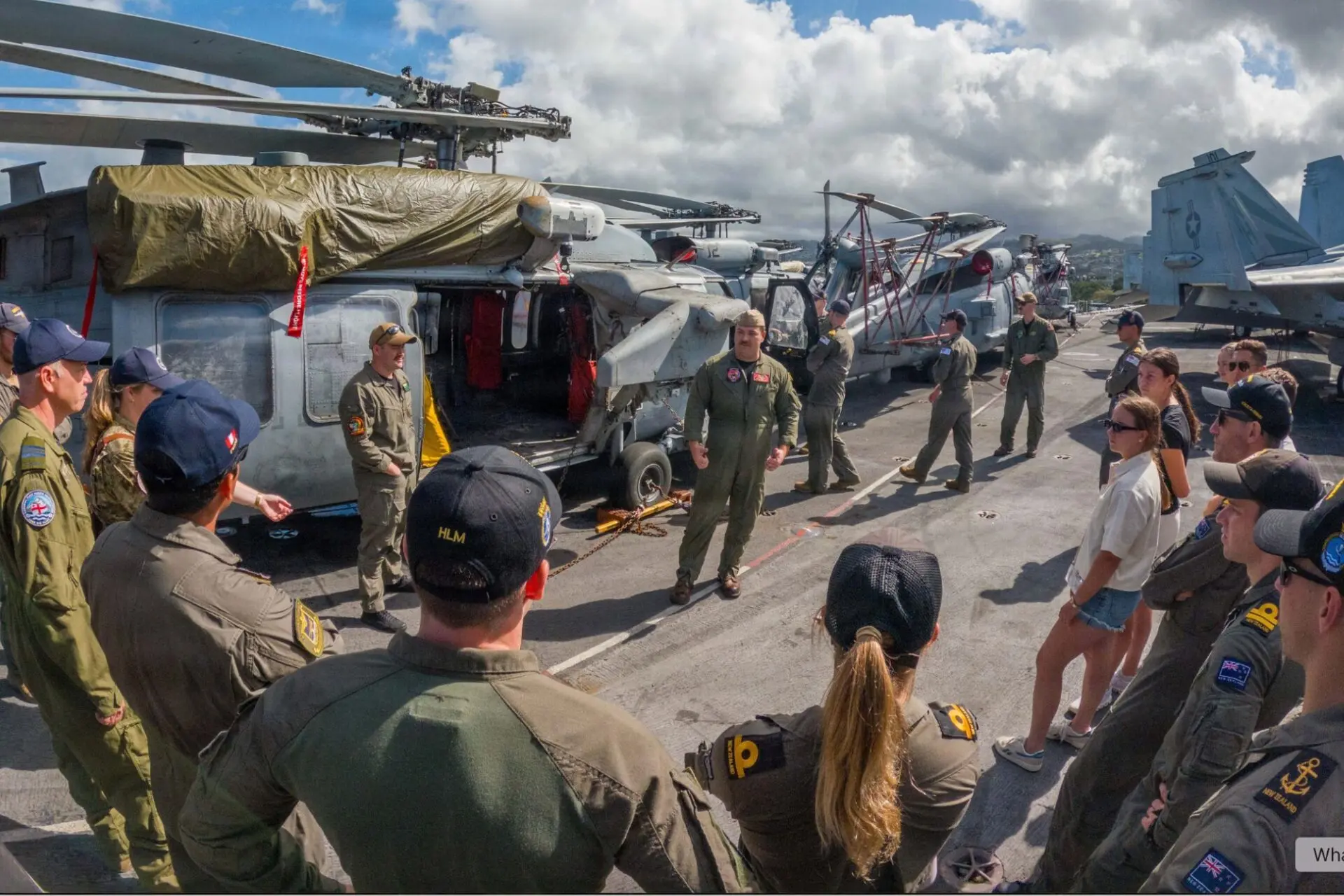 Crew members in flight suits stand on an aircraft carrier deck beside parked helicopters and aircraft structures in the background