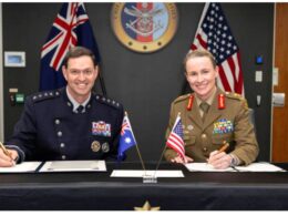 Two uniformed officersone in a dark navy dress uniform and the other in a khaki army uniformsmile while signing documents at a formal desk with Australian and American flags in front and an official seal on the wall behind them