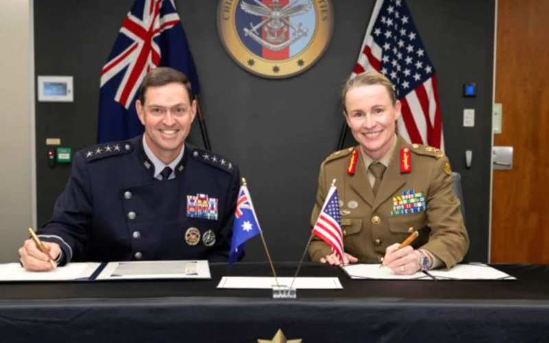 Two uniformed officersone in a dark navy dress uniform and the other in a khaki army uniformsmile while signing documents at a formal desk with Australian and American flags in front and an official seal on the wall behind them