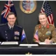 Two uniformed officersone in a dark navy dress uniform and the other in a khaki army uniformsmile while signing documents at a formal desk with Australian and American flags in front and an official seal on the wall behind them