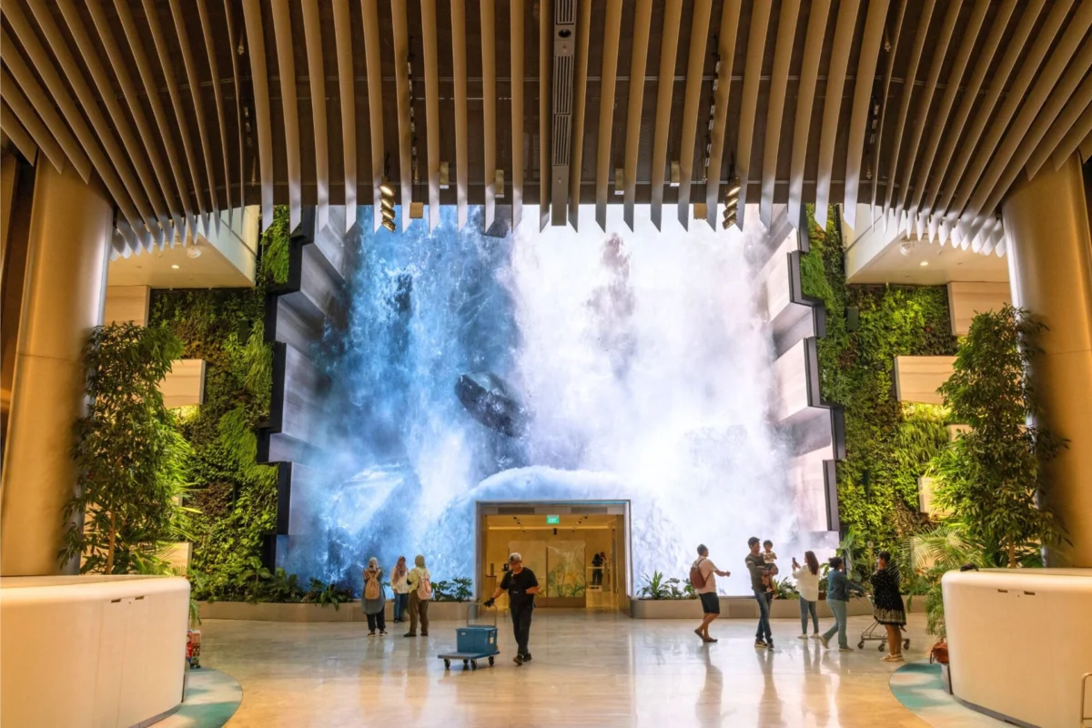 Indoor atrium with a large waterfall feature at the far end surrounded by plants and visitors walking around