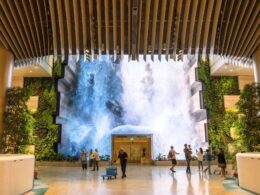 Indoor atrium with a large waterfall feature at the far end surrounded by plants and visitors walking around