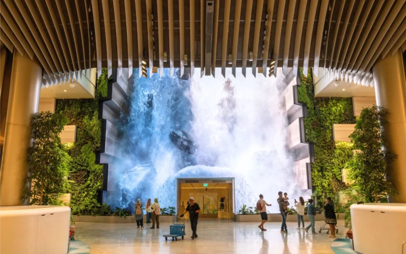 Indoor atrium with a large waterfall feature at the far end surrounded by plants and visitors walking around