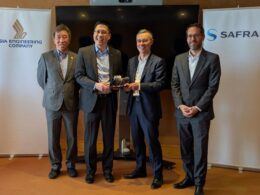Four men in business suits stand side by side at a bannered event two of them handing a small trophy to each other as a presentation moment