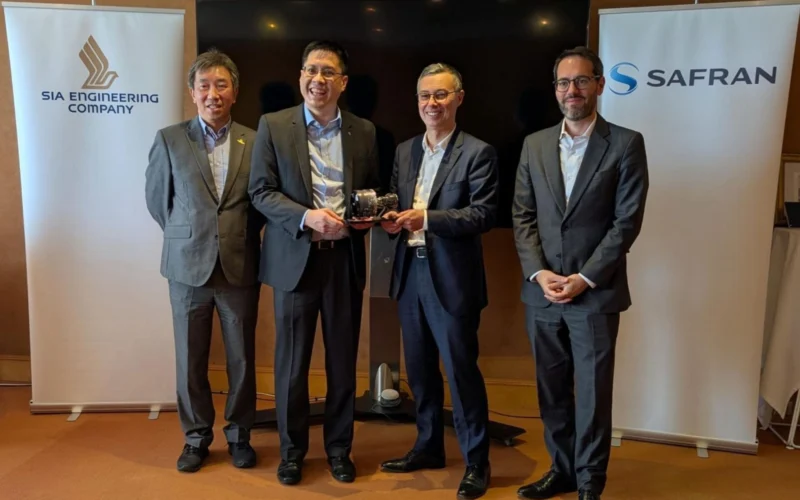 Four men in business suits stand side by side at a bannered event two of them handing a small trophy to each other as a presentation moment