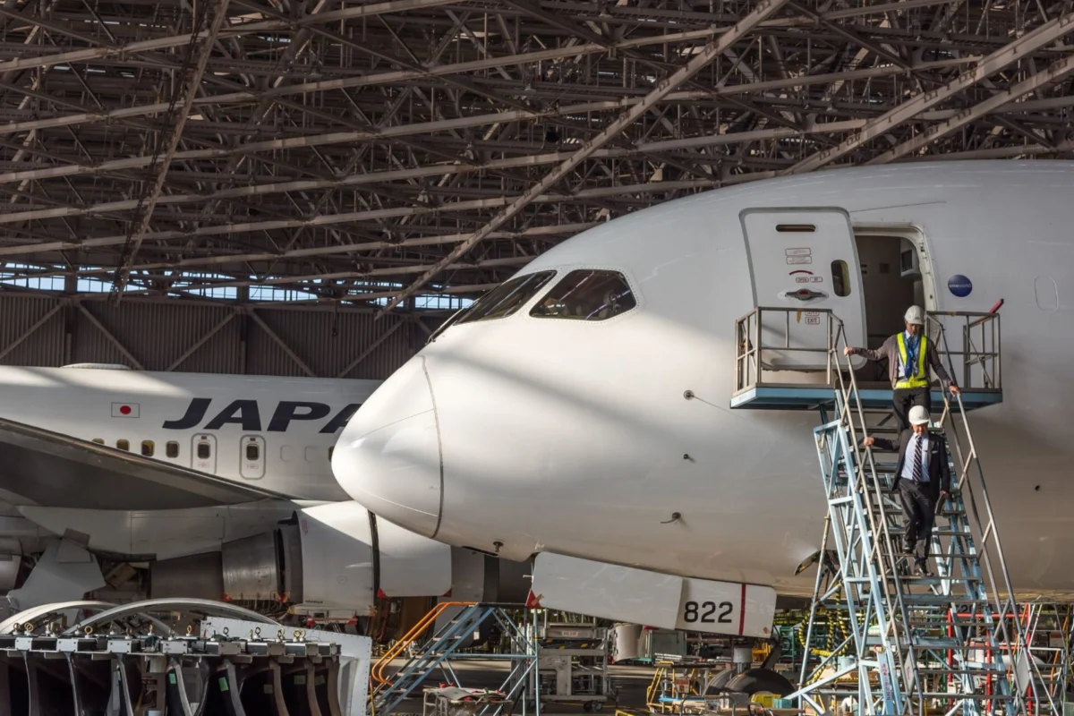 Japan Airlines jet under a metal hangar roof with maintenance workers on a scaffold beside the open door of the forward fuselage